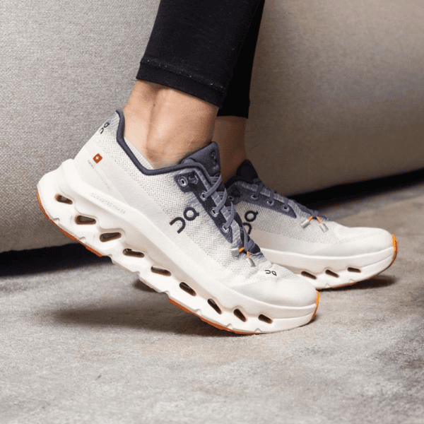 Pair of gray and white running shoes with orange accents worn by someone standing on a carpeted floor.
