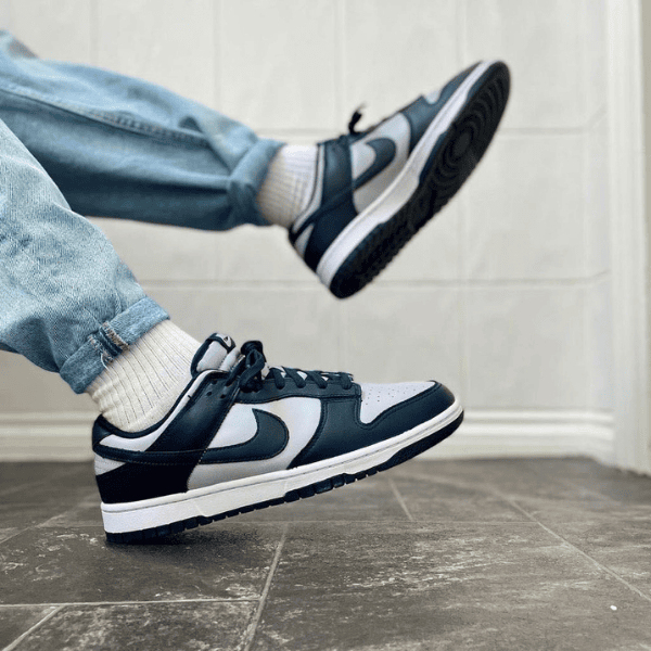 Person wearing light-wash jeans and white socks kicks a navy and white sneaker mid-air on a tiled floor, showing the side of the shoe.