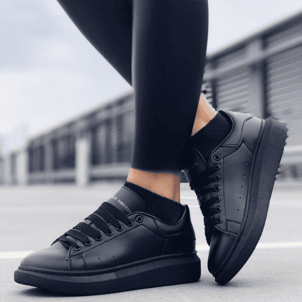 Black sneakers with matching black leggings worn on a paved outdoor surface, urban backdrop in the distance.