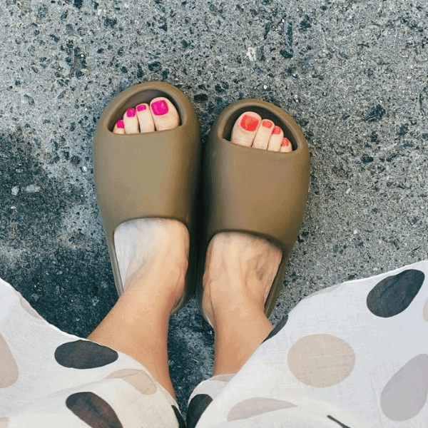 Top-down view of feet wearing olive-green slide sandals on a concrete surface, showing pink and orange pedicure nails.