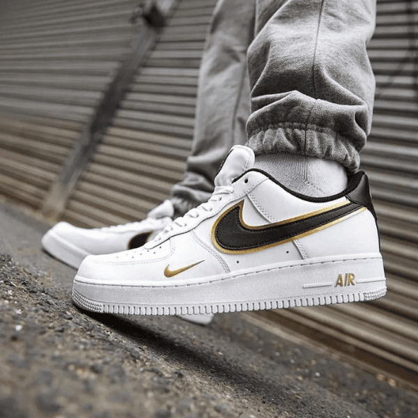 White Nike Air Force 1 sneakers with black and gold swoosh, worn with gray joggers, standing on a curb.