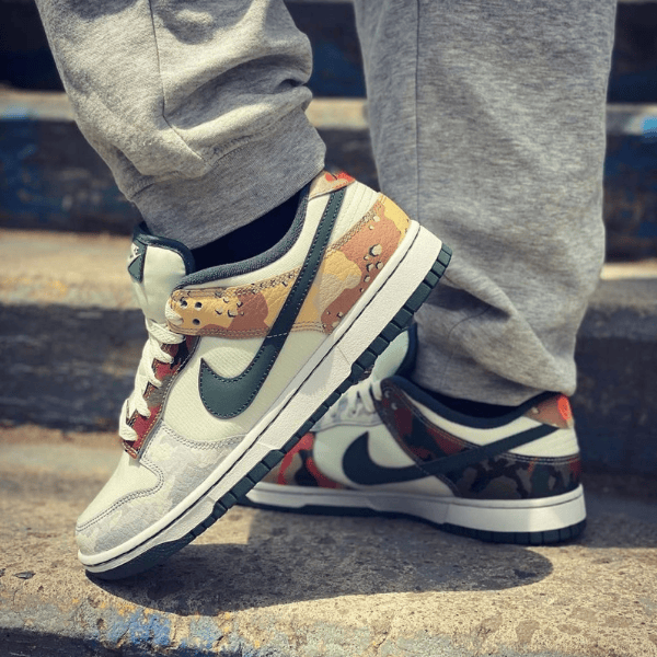 Close-up of white and gray Nike sneakers with camouflage panels and a green swoosh on concrete stairs with gray joggers nearby.