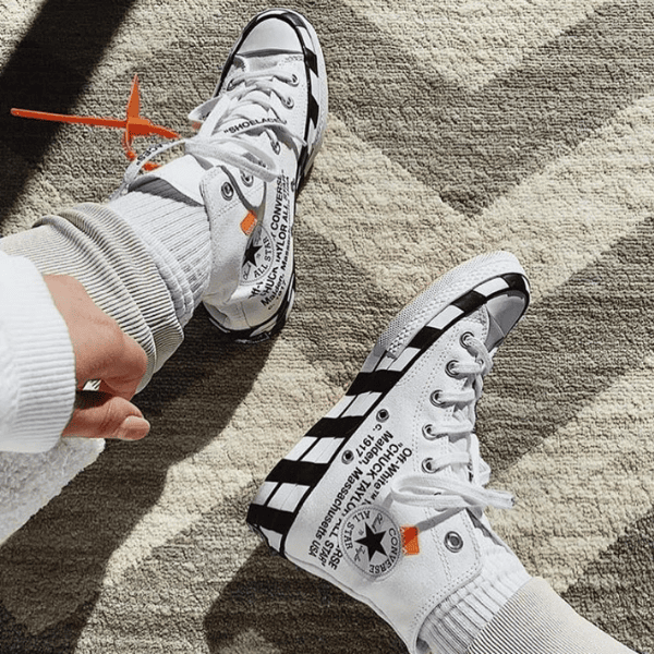 Two white sneakers with black stripes on a beige rug, laces tied, orange tag on the left shoe with text visible on the sides of both sneakers.