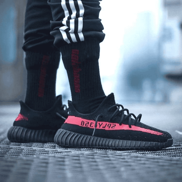 Close-up of a person wearing black and red sneakers with a pink stripe on a metal grate, urban setting in the background.