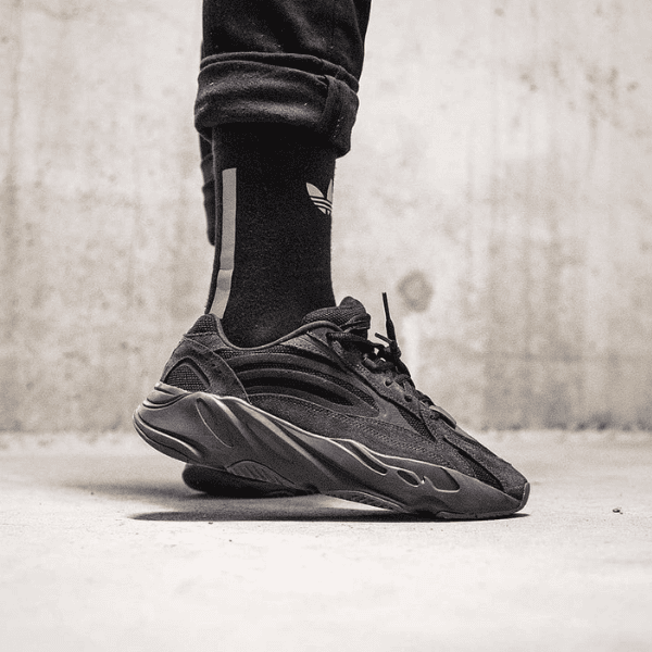 Black chunky sneaker on a foot wearing a black sock with a white logo, standing on a concrete floor.