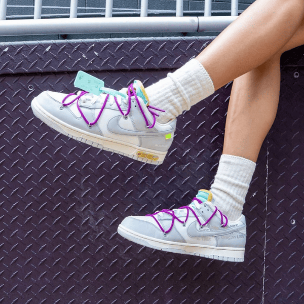 Person wearing light gray sneakers with bright pink laces and white socks, legs resting on a purple diamond-plate surface with tags on the shoes nearby.