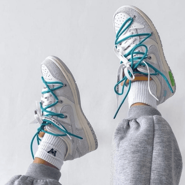 Two light gray sneakers with teal laces and white socks, shown raised against a light background (casual athletic look).