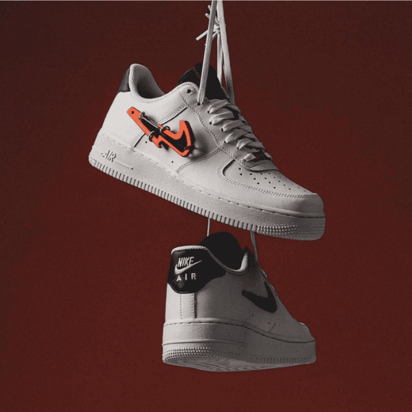 Two white Nike Air sneakers hanging by their laces against a dark red backdrop, with an orange tag on the top shoe.