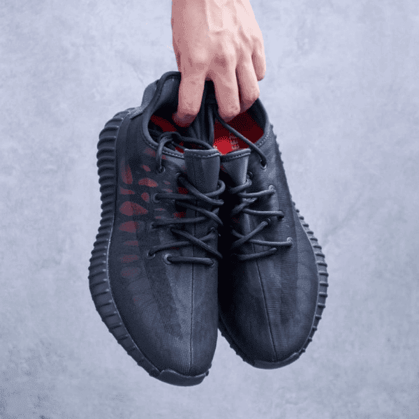 A person holds two black lace-up sneakers by the top, showing the side and red inner lining against a gray background.