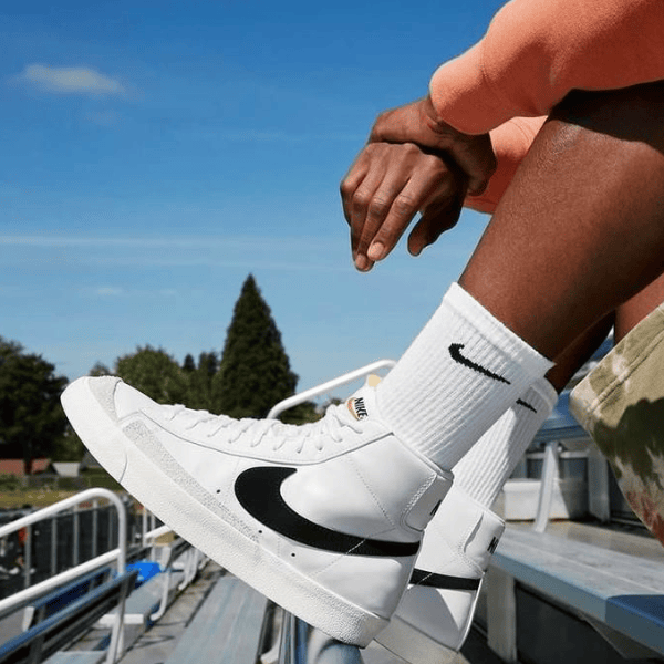 White Nike high-top sneakers with a black swoosh, worn with white socks, resting on bleachers outdoors on a sunny day.