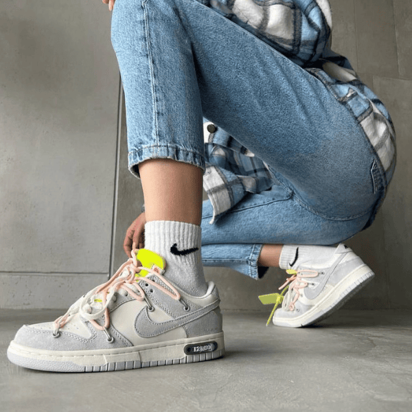 Person crouching and tying light gray Nike sneakers with pink laces, white socks, and light-wash jeans in a casual indoor setting