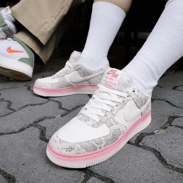 Close-up of white Nike Air sneakers with pink midsole worn with white socks, standing on a paved surface.