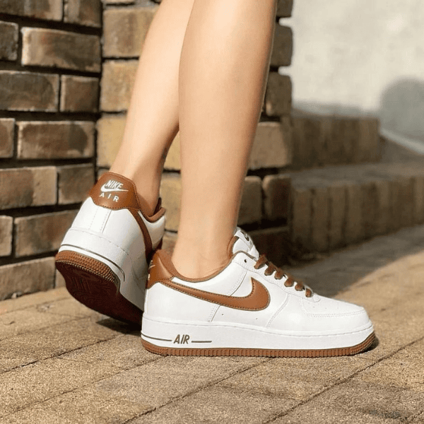 White Nike Air sneakers with brown accents and swoosh, worn by a person standing on a brick walkway near steps.