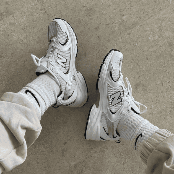Two white New Balance sneakers worn with light socks and rolled-up pants, resting on a concrete floor.