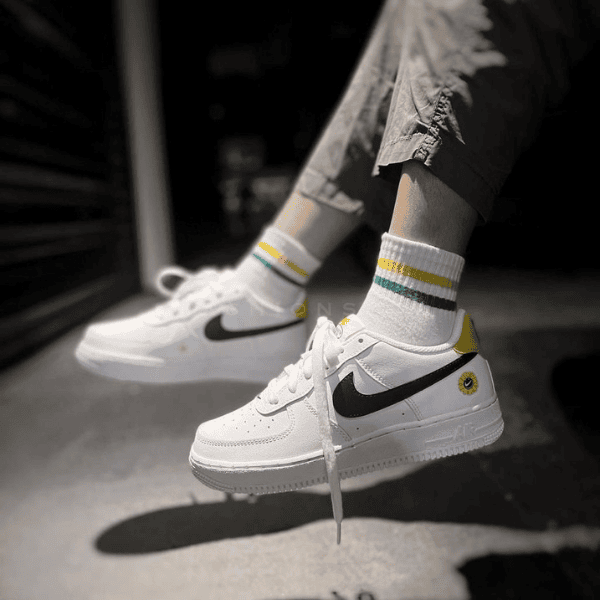 White Nike sneakers with black swoosh and yellow heel tab, worn with socks that have green and yellow stripes, shown mid-air as the person walks or jumps.