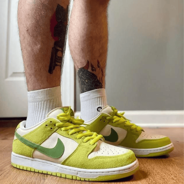 Person wearing lime-green Nike-style sneakers with white panels and lime laces, standing on a wooden floor with tattoos visible on the legs.