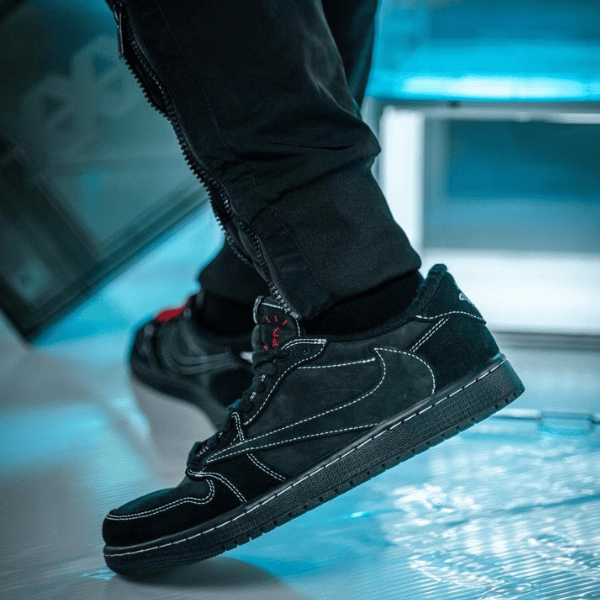 Close-up of a black suede sneaker with white stitching, worn by a person stepping forward on a blue-lit floor.