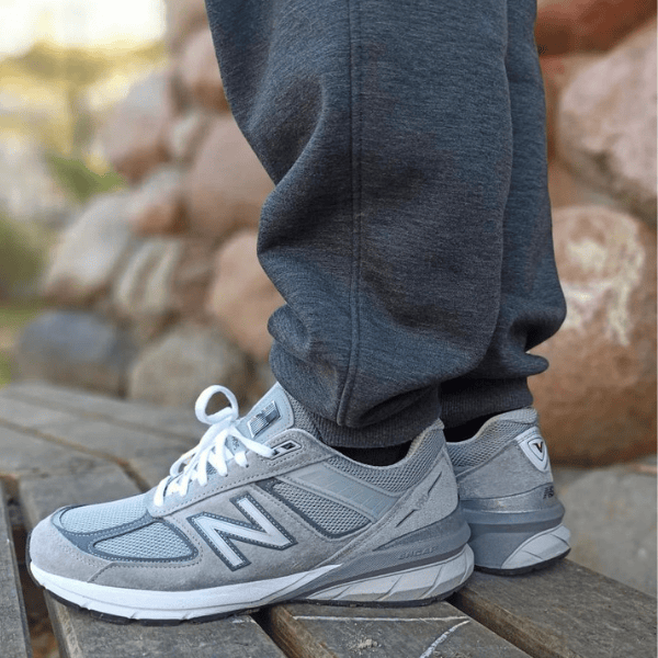 Gray New Balance running shoes with white laces on a person standing on a wooden surface, with rocks in the background.