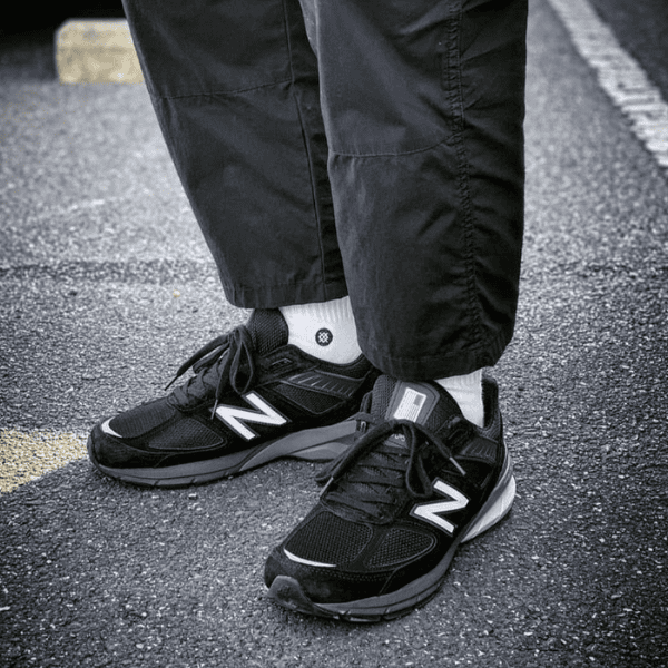 Pair of black New Balance sneakers with white socks standing on a street or parking lot surface.