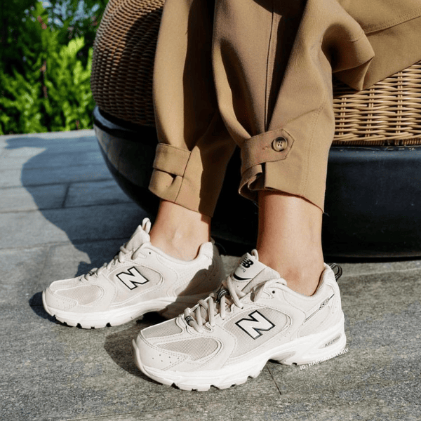 Person wearing beige pants and cream New Balance sneakers standing on a stone patio with a wicker chair nearby