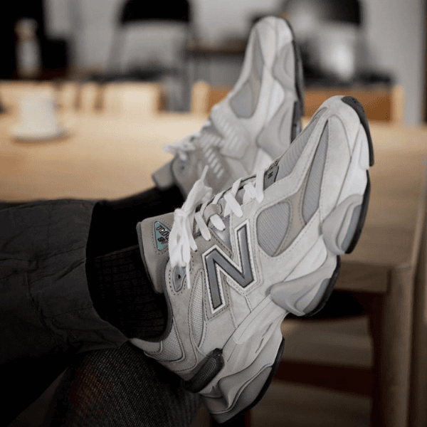Pair of white and gray New Balance sneakers resting on someone’s feet in an indoor casual setting with a wooden table in the background.