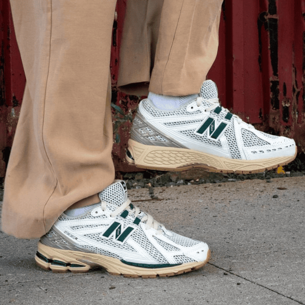 Pair of white and gray New Balance running shoes with green NB logo, worn with tan pants on a sidewalk.