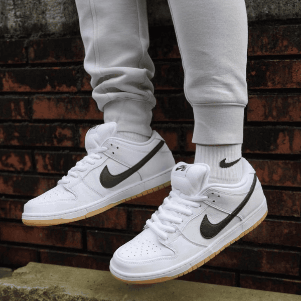 Person wearing white Nike sneakers with black swoosh and gum soles, standing on a ledge in gray joggers by a brick wall