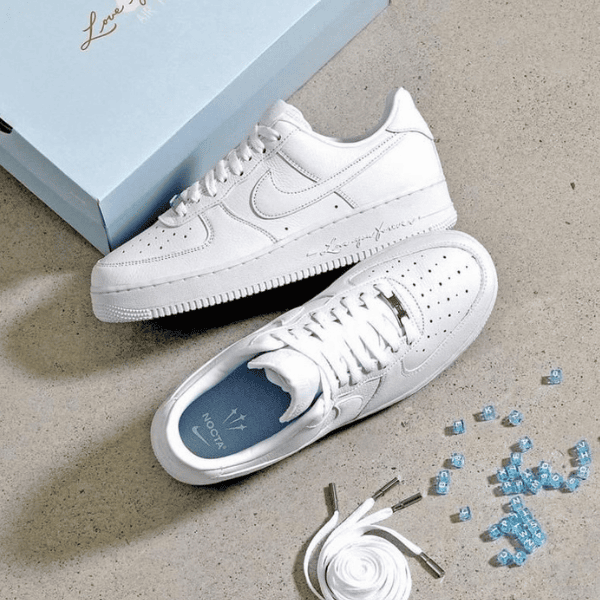 White sneakers with perforated toe cap placed on a light gray floor next to a pale blue shoebox; laces untied, blue insole visible, and small blue decorative beads with a coiled white shoelace nearby.