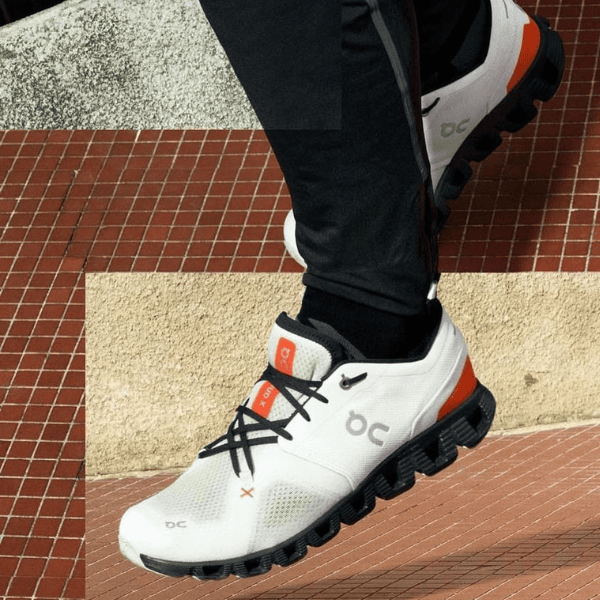 White athletic running shoe with black laces and orange details, stepping up a red-tiled staircase; visible OC logo on the side.