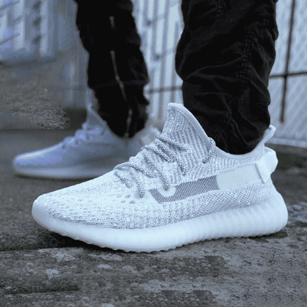 Glittery white knit sneakers on a person standing on a concrete surface, wearing black pants, side view showcasing the shoe's textured upper and thick sole.