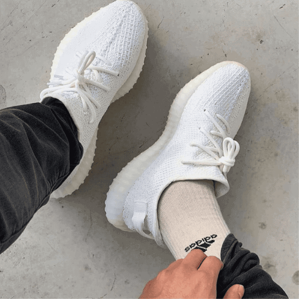 Top-down view of white knit sneakers with chunky off-white soles, paired with beige Adidas-branded socks on a gray floor.
