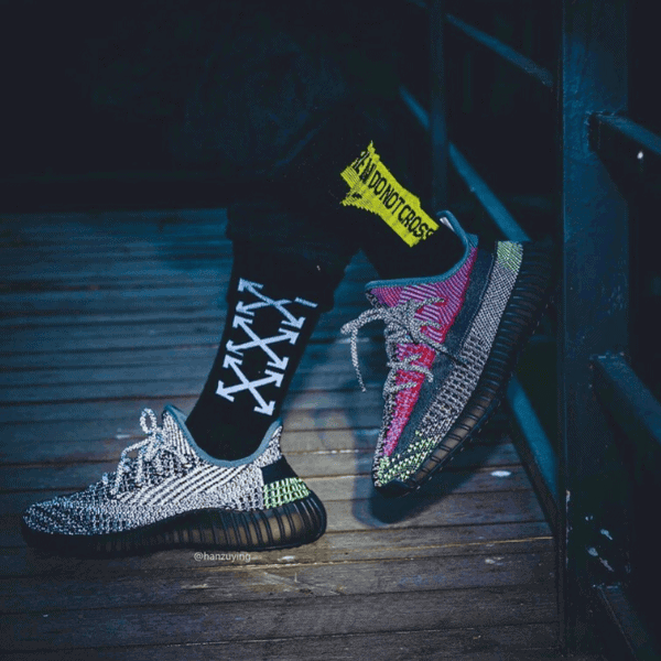 Pair of sporty sneakers on a wooden deck, one foot in a black sock with white arrows and a yellow taped label nearby.