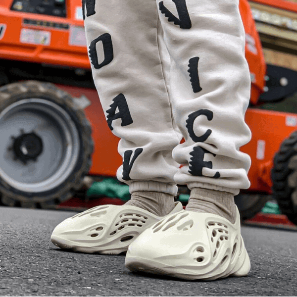 Person wearing light gray joggers with black scattered letters, standing in white sneakers on pavement, orange machinery in the background.