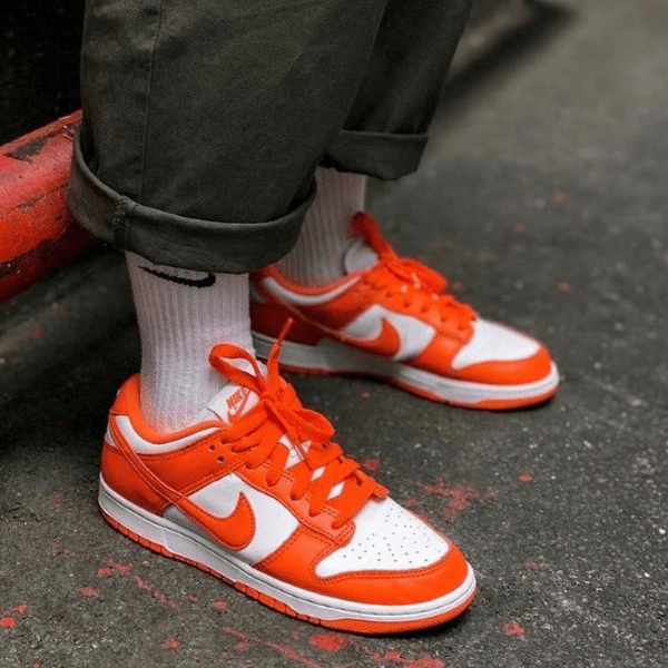 Close-up of a person wearing white crew socks and orange-and-white Nike sneakers standing on a dark pavement.