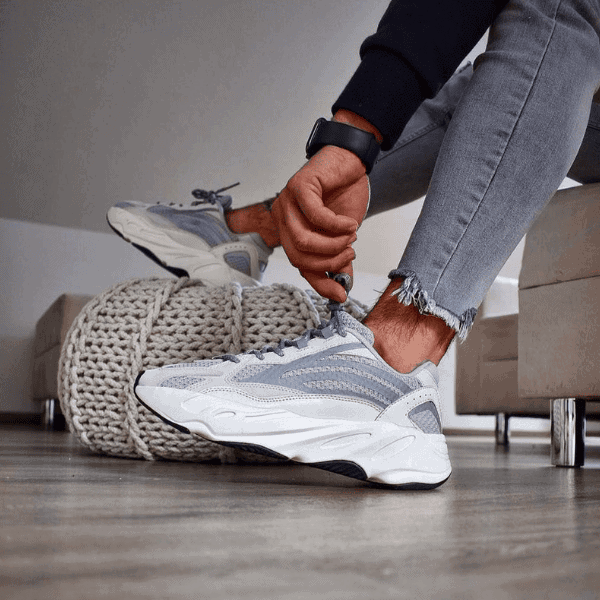 Person tying the laces of gray and white athletic sneakers, seated indoors with a chunky knit pouf nearby.