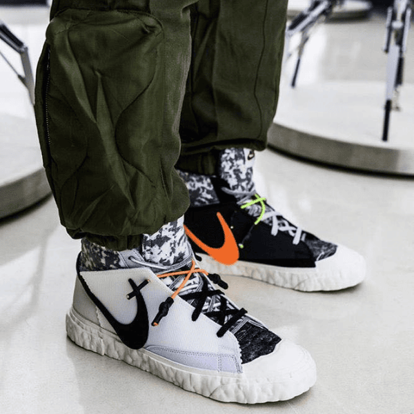 Close-up of two Nike sneakers: the foreground white high-tops with black swoosh and orange laces, and a black pair with neon green laces in the background, worn with camo socks and olive cargo pants.