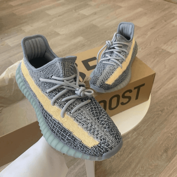 Gray knit sneakers with a pale yellow side stripe and light teal sole, resting on a BOOST box in a home setting.