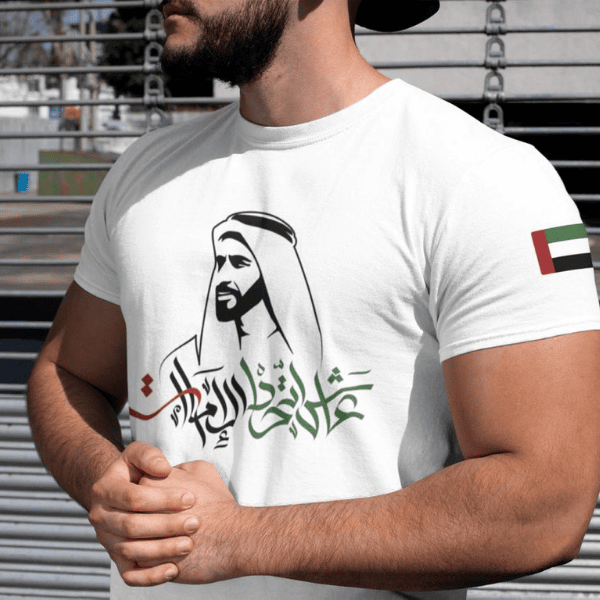 Man in a white T-shirt with green Arabic calligraphy and a UAE flag patch on the sleeve, standing with hands clasped.