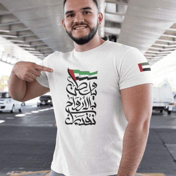 Man in a white t-shirt pointing to bold Arabic calligraphy text on the chest, with UAE flag colors above it and a flag on the sleeve, in an outdoor setting.