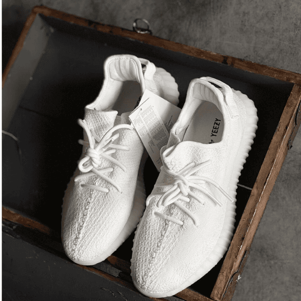 Pair of white knit sneakers with laces, placed in an open wooden crate; Yeezy branding visible inside.