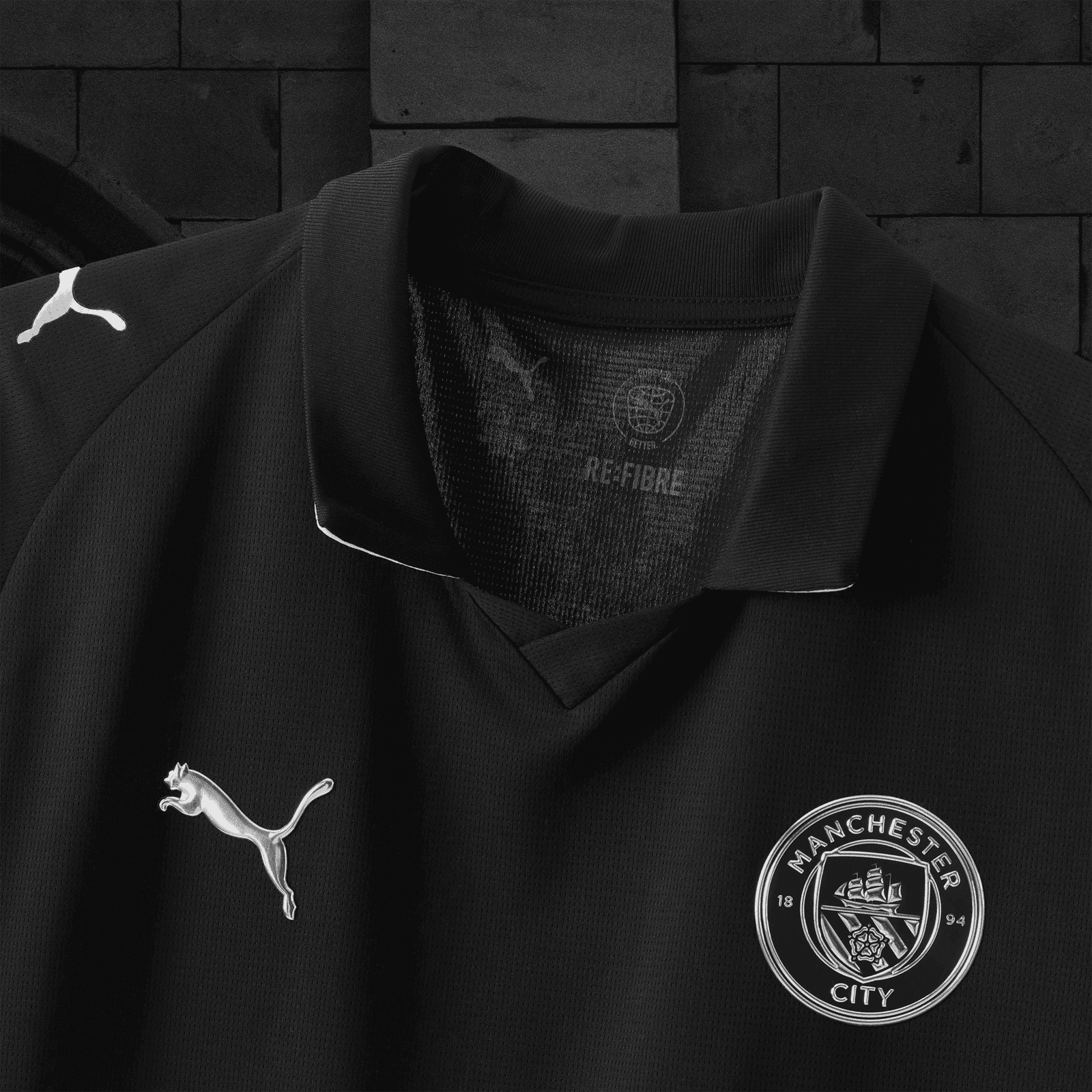 V3-1 | AE JERSEY Close-up of a black Puma soccer jersey showing the white Puma logo near the shoulder and the Manchester City club crest on the chest, with a textured fabric background.