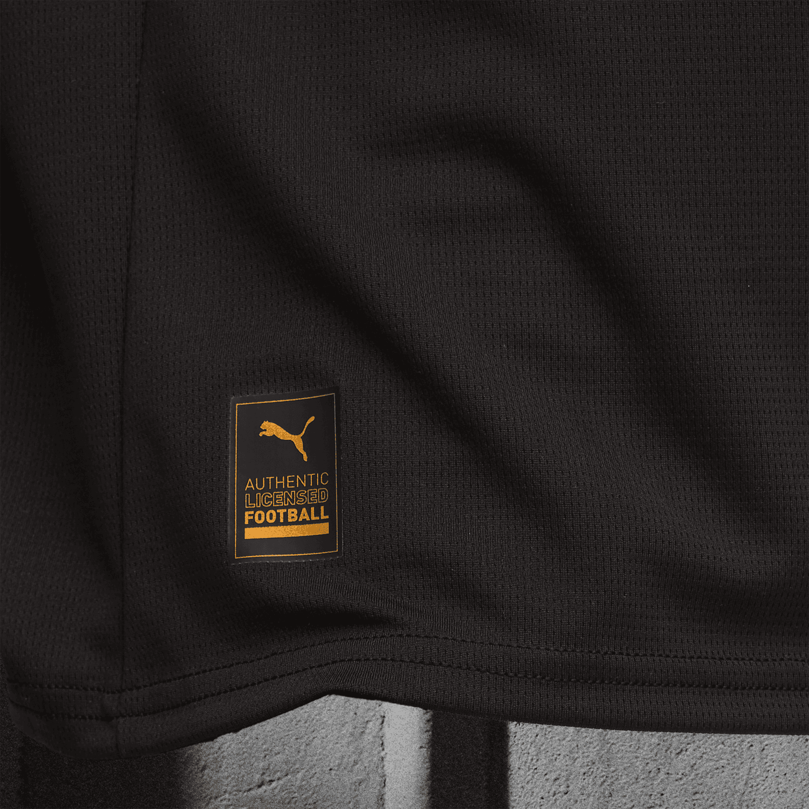 V5-1 | AE JERSEY Close-up of black textured sports fabric with an orange Puma label that says 'AUTHENTIC LICENSED FOOTBALL'.