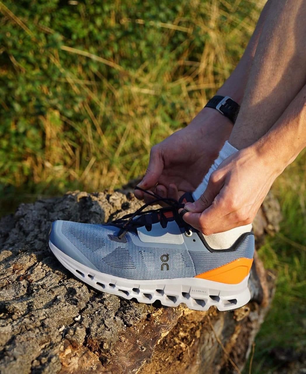 WhatsAppImage2024-11-21at12.03.31 | AE JERSEY Person tying the laces of a light blue running shoe with an orange heel on a rocky surface outdoors