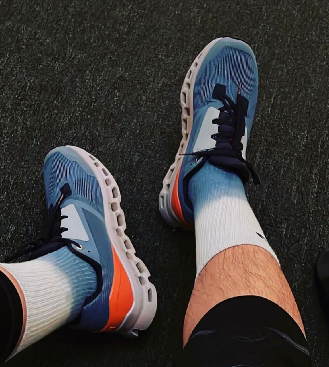 WhatsAppImage2024-11-21at12.03.42 | AE JERSEY Person wearing blue athletic sneakers with white soles and orange accents, standing on a dark textured surface in white socks.