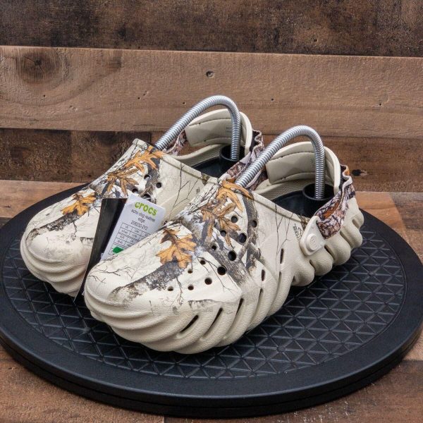 Pair of beige camouflage Crocs with ventilation holes, displayed on a black circular platform, tags attached to the shoe.