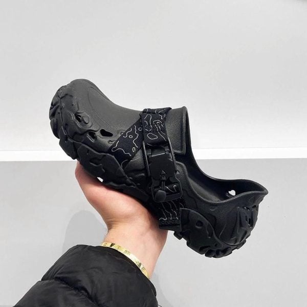 Black chunky slip-on clog with a patterned strap, held in a hand against a light background.