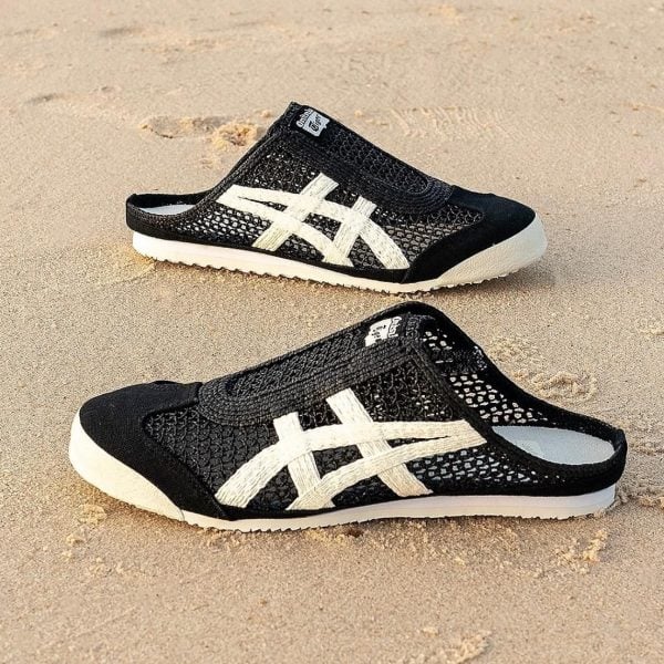 Black mesh slip-on shoes with white crisscross stripes on sandy beach surface.