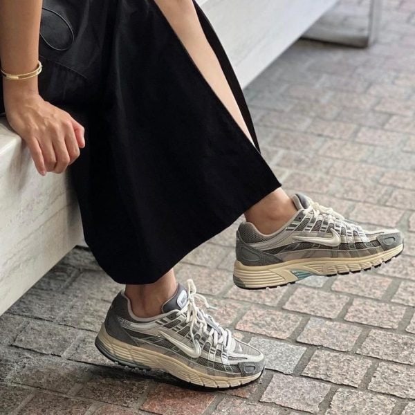 Person in a black dress sits with legs extended, wearing gray running sneakers on a brick sidewalk.