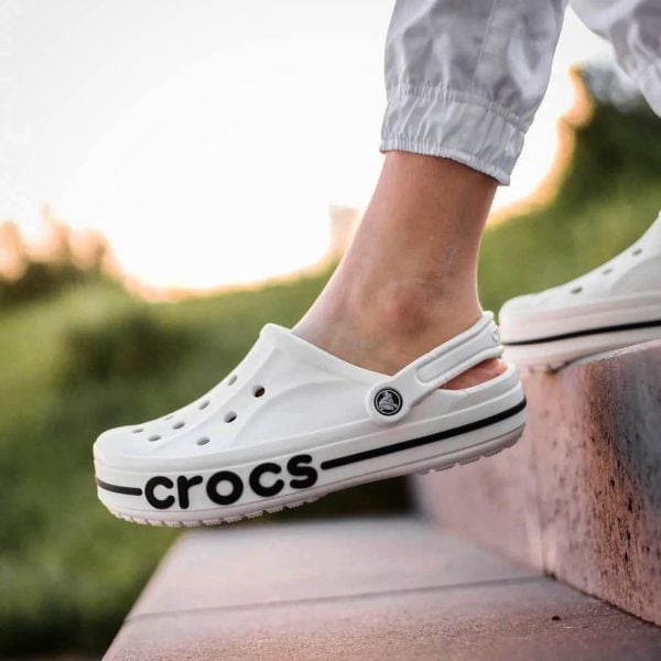 White Crocs clog with bold black 'CROCS' lettering along the sole, worn by a person stepping onto a concrete step outdoors.