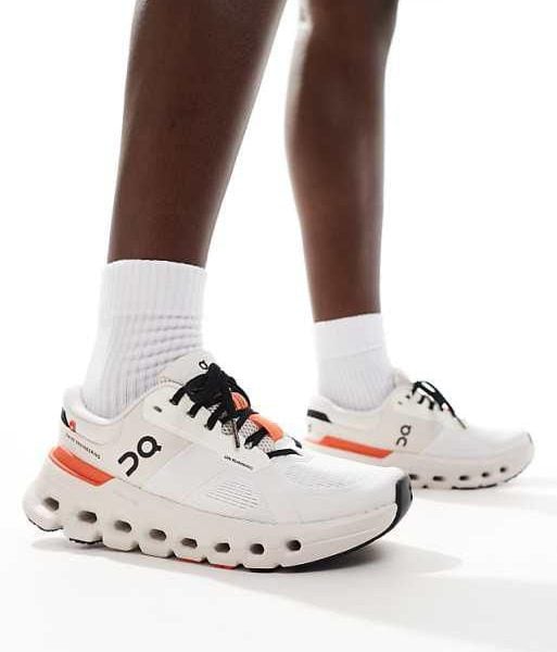 Person wearing white running shoes with a chunky sole and orange heel accents, standing with white socks.